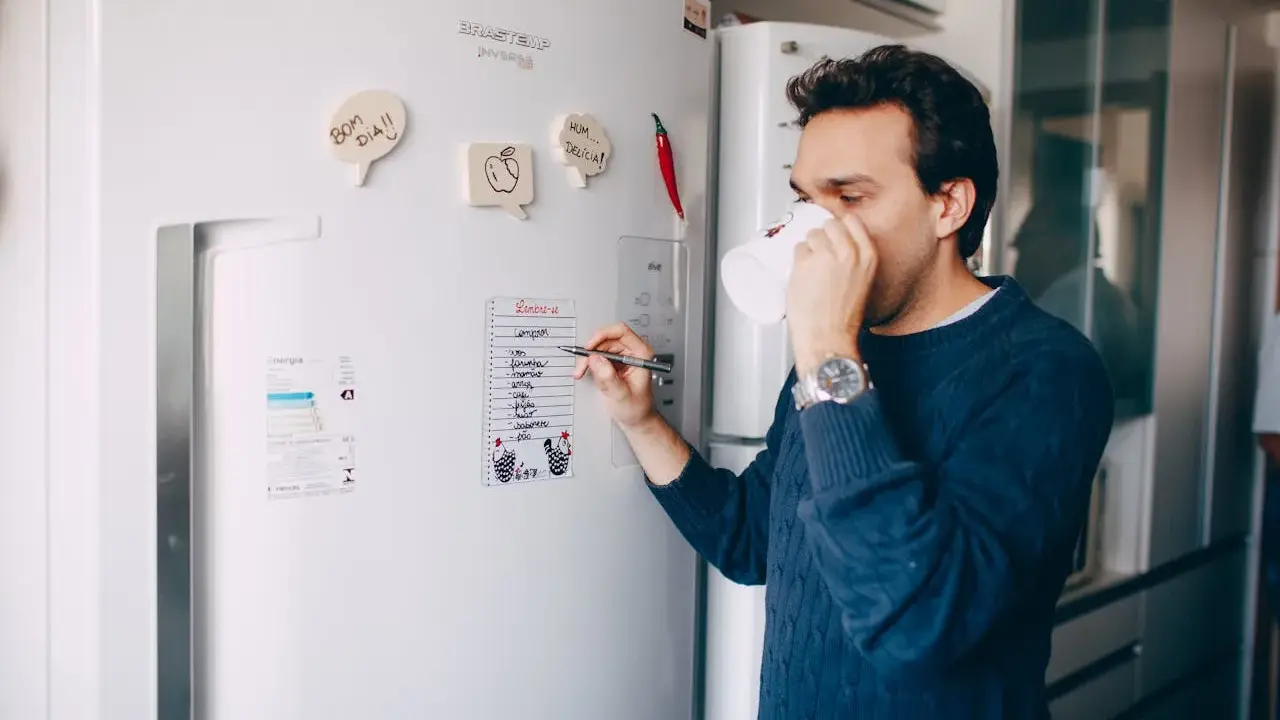 Mężczyzna z kubkiem kawy pisze listę zakupów na lodówce no frost.