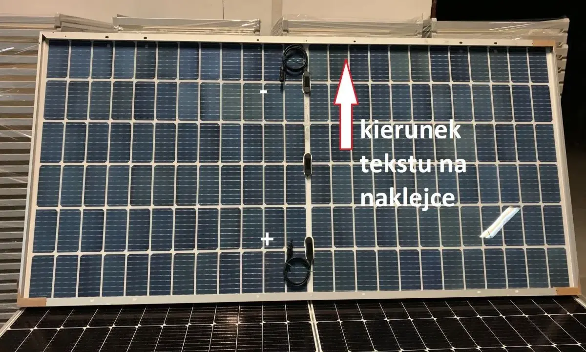 Panel fotowoltaiczny z oznaczeniami plus i minus. Strzałka wskazuje kierunek tekstu na naklejce.