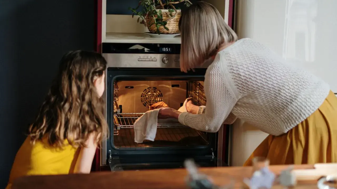 Mama i córka przyglądają się pierwszemu użyciu piekarnika. Kobieta wyjmuje z niego upieczone ciasto.