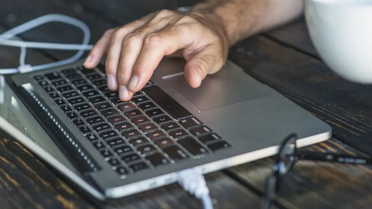 Dłoń na klawiaturze laptopa, przygotowująca się do twardego resetu. Obok biała filiżanka i okulary.