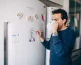 Mężczyzna z kubkiem kawy pisze listę zakupów na lodówce no frost.