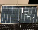 Panel fotowoltaiczny z oznaczeniami plus i minus. Strzałka wskazuje kierunek tekstu na naklejce.