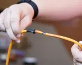 Dłonie trzymają przecięty pomarańczowy kabel, odsłaniając niebieskie żyły. Pokazuje to, jak połączyć przecięty kabel w ścianie.
