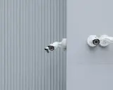 Montaż kamery na elewacji budynku. Dwie białe kamery monitoringu zamontowane na szarej ścianie, jedna na tle pionowych paneli.