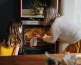 Mama i córka przyglądają się pierwszemu użyciu piekarnika. Kobieta wyjmuje z niego upieczone ciasto.