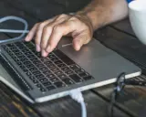 Dłoń na klawiaturze laptopa, przygotowująca się do twardego resetu. Obok biała filiżanka i okulary.