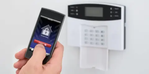 Dłoń trzyma smartfon z aplikacją alarmową, obok panel alarmowy. Idealny jaki alarm do mieszkania w bloku.