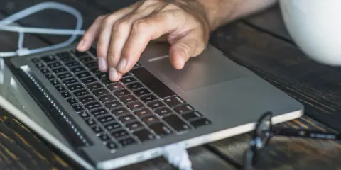 Dłoń na klawiaturze laptopa, przygotowująca się do twardego resetu. Obok biała filiżanka i okulary.