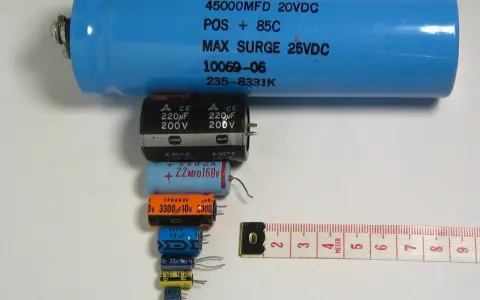 Różne kondensatory elektrolityczne, od dużego niebieskiego po małe czarne i kolorowe, ułożone obok miarki. Budowa kondensatora elektrolitycznego jest tu widoczna w całej okazałości.