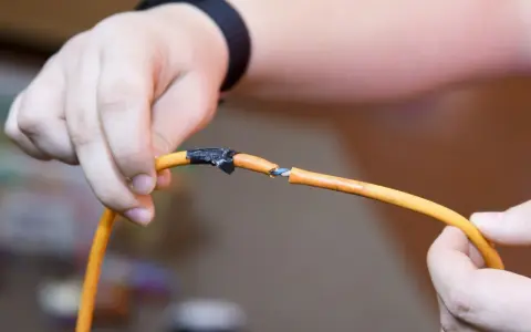 Dłonie trzymają przecięty pomarańczowy kabel, odsłaniając niebieskie żyły. Pokazuje to, jak połączyć przecięty kabel w ścianie.