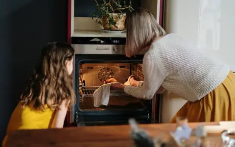 Mama i córka przyglądają się pierwszemu użyciu piekarnika. Kobieta wyjmuje z niego upieczone ciasto.