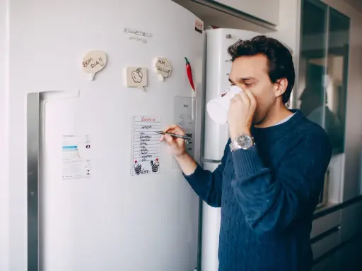 Mężczyzna z kubkiem kawy pisze listę zakupów na lodówce no frost.