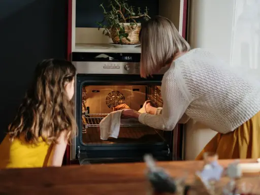 Mama i córka przyglądają się pierwszemu użyciu piekarnika. Kobieta wyjmuje z niego upieczone ciasto.