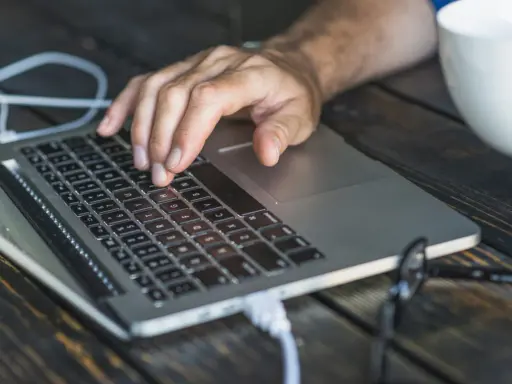 Dłoń na klawiaturze laptopa, przygotowująca się do twardego resetu. Obok biała filiżanka i okulary.