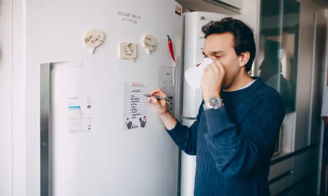 Mężczyzna z kubkiem kawy pisze listę zakupów na lodówce no frost.