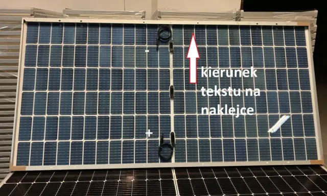 Panel fotowoltaiczny z oznaczeniami plus i minus. Strzałka wskazuje kierunek tekstu na naklejce.