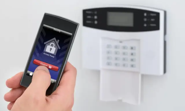 Dłoń trzyma smartfon z aplikacją alarmową, obok panel alarmowy. Idealny jaki alarm do mieszkania w bloku.