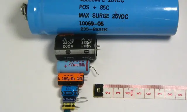 Różne kondensatory elektrolityczne, od dużego niebieskiego po małe czarne i kolorowe, ułożone obok miarki. Budowa kondensatora elektrolitycznego jest tu widoczna w całej okazałości.
