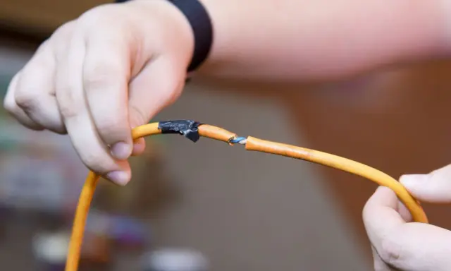 Dłonie trzymają przecięty pomarańczowy kabel, odsłaniając niebieskie żyły. Pokazuje to, jak połączyć przecięty kabel w ścianie.