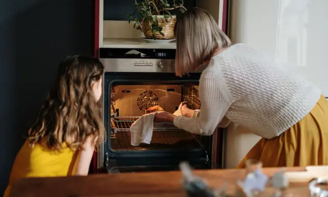 Mama i córka przyglądają się pierwszemu użyciu piekarnika. Kobieta wyjmuje z niego upieczone ciasto.