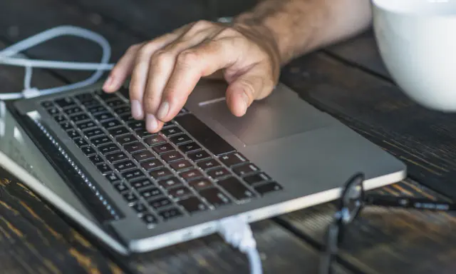 Dłoń na klawiaturze laptopa, przygotowująca się do twardego resetu. Obok biała filiżanka i okulary.