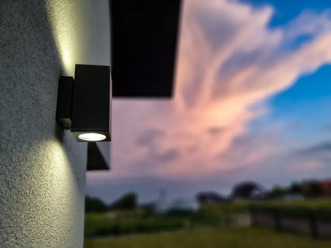 Nowoczesny montaż lampy na elewacji oświetla ścianę. W tle zach&oacute;d słońca z r&oacute;żowymi chmurami.