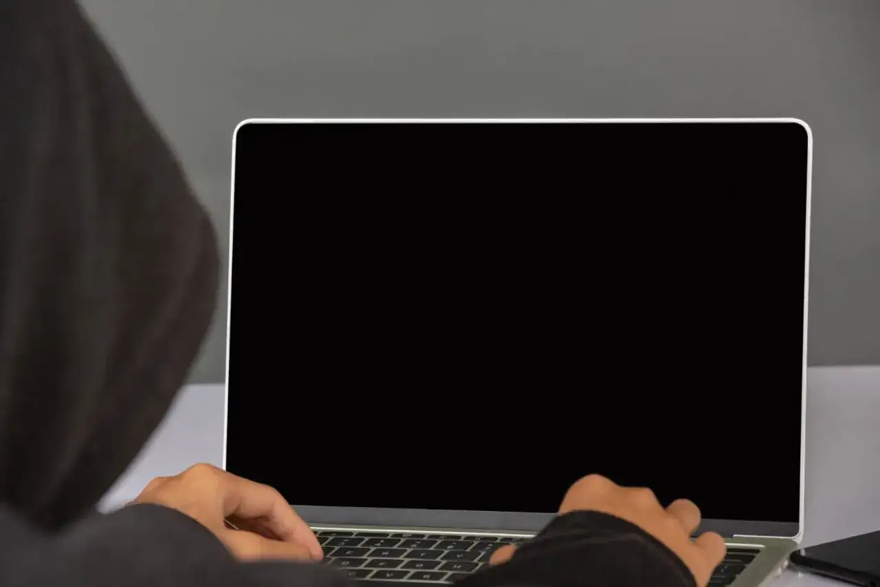 Osoba w kapturze pracuje na laptopie z czarnym ekranem.