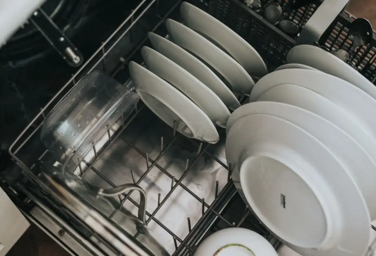 Wnętrze zmywarki z talerzami i plastikowym pojemnikiem. Białe naczynia są ułożone na p&oacute;łce. Widoczne są objawy za dużo soli w zmywarce: biały nalot na naczyniach.
